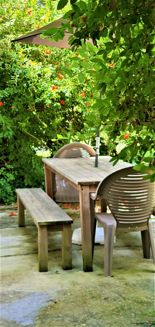 Beschatteter Essbereich im Freien bei Le Manoir Lacustre mit Holztisch, Bank und gestreiften Stühlen umgeben von üppigem grünem Garten
