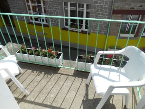 Balcon ensoleillé à L'Abri Des Vents avec chaises blanches, jardinières fleuries et balustrade verte surplombant Saint-Jean-le-Thomas