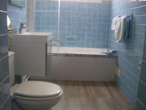 Salle de bains moderne aux murs carrelés bleus, sanitaires blancs, baignoire et sol en bois à l'Abri Des Vents