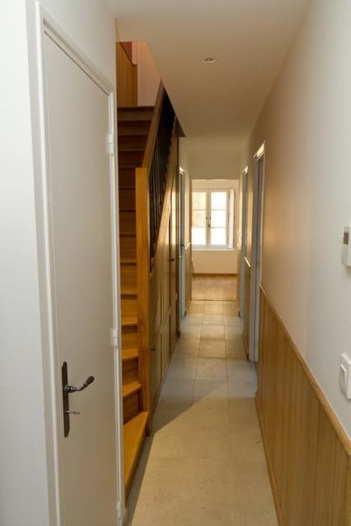 Long couloir du gîte Le Fassardy avec escalier en bois, portes blanches et lumière naturelle de la fenêtre