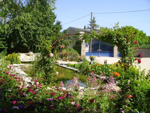 Vue bucolique du jardin de la maison d'hôtes La Chabanaise en Marais Poitevin : fleurs colorées en pleine floraison, petit bassin aquatique et portail bleu typique sous un ciel ensoleillé.