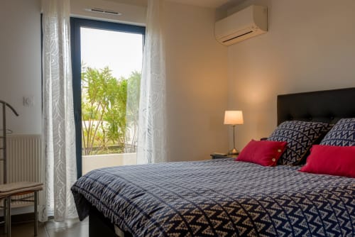 Chambre d'appartement en Provence avec vue sur jardin, literie à motifs bleus, oreillers rouges, climatisation moderne et accès au balcon