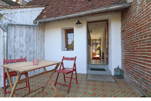 Terrasse de la maison d'hôtes La Dragonne à Bellême avec table en bois, chaises rouges et bâtiment en brique avec vue sur le jardin