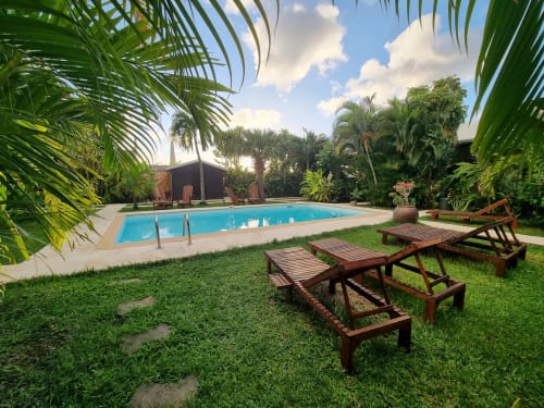 Área de piscina tropical en Gites Kaladja en Port-Louis con tumbonas de madera, exuberantes jardines de palmeras y bungaló con techo de paja