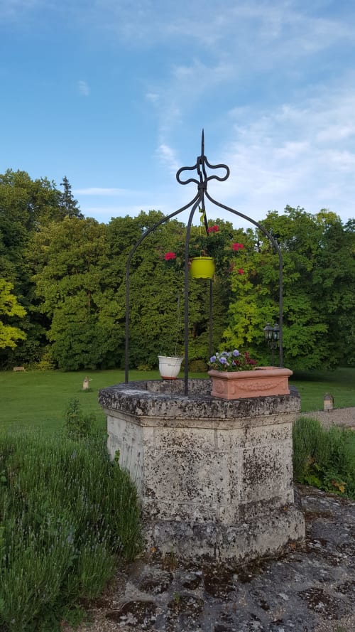 Ornate garden well with black iron cross at Château de Ré, Le Petit Pressigny, surrounded by blooming flowers and lush trees
