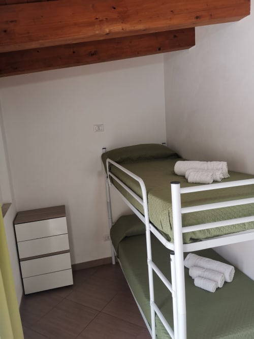 Cozy bunk bedroom with white metal bed frame, green mattresses, rolled towels, wooden beams, and wall outlet at Casa Vacanze