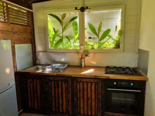 Compact kitchen with wooden cabinets, gas stove, sink, and window overlooking tropical plants at Gites Kaladja apartment in