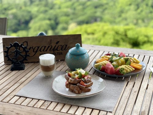 Repas en plein air à Organica Yoga Shelter : cappuccino, bruschetta, légumes grillés et fruits tropicaux sur terrasse en bois avec vue océan