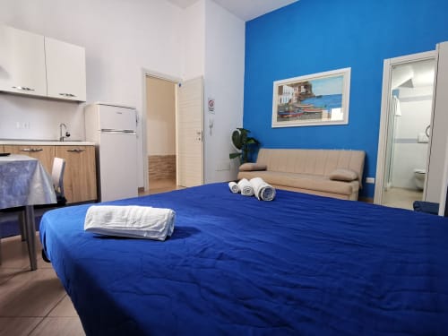 Bright studio apartment with blue accent wall, queen bed with blue comforter, kitchenette, and beige sofa at Casa Vacanze Del