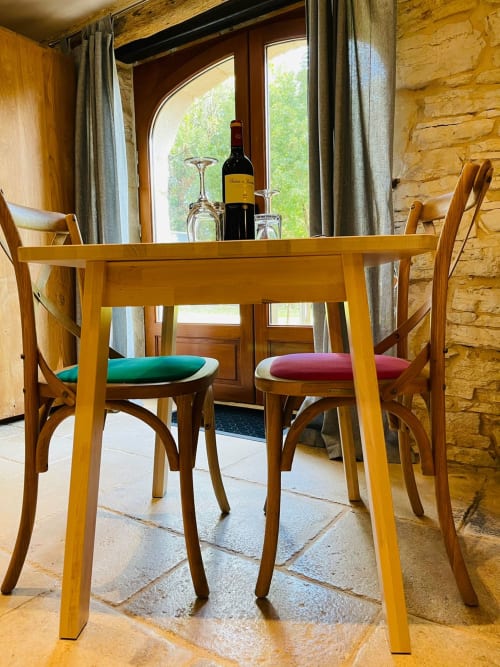 Table de salle à manger en bois avec coussins de chaise colorés au gîte de Gindou, bouteille de vin et verres sur la table, murs en pierre et jardin
