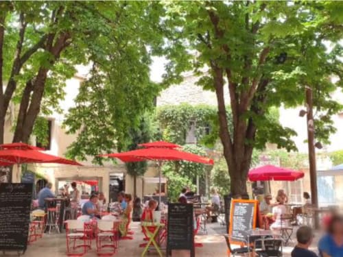 Outdoor courtyard at Provence Km28 in Gordes with red umbrellas, shaded by trees, featuring dining seating and cafe ambiance.