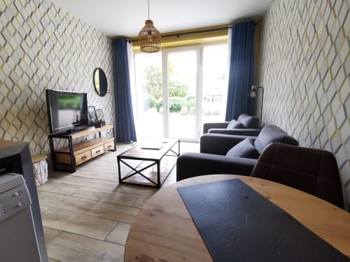 Salon moderne à La Cour d'Hortense avec canapé noir, table en bois, TV et vue sur le jardin par les portes-fenêtres