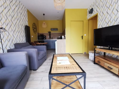 Salon cosy à La Cour d'Hortense avec fauteuils bleus, murs jaunes, cuisine, TV écran plat et motifs