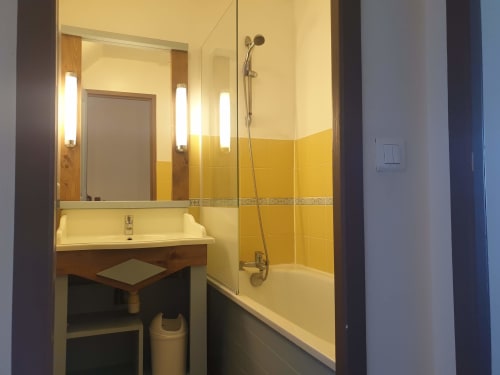 Salle de bain moderne de l'appartement Locmaria avec carrelage jaune, meuble vasque, baignoire avec douche et miroir lumineux
