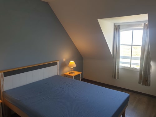 Chambre mansardée moderne avec lit bleu, tête de lit blanche, table de nuit en bois avec lampe et fenêtre de toit offrant une vue sur Belle Île