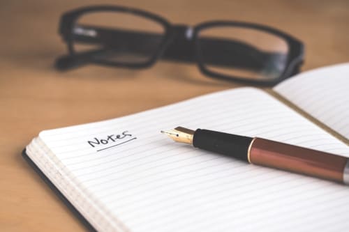 Open notebook with 'Notes' written on lined page, fountain pen, and reading glasses on wooden desk at Domaine de Blanvatel