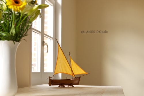 Pièce baignée de soleil avec maquette de voilier jaune, vase blanc de tournesols, et texte 'Palaises d'Opale' à La ferme de l'Ostrevent