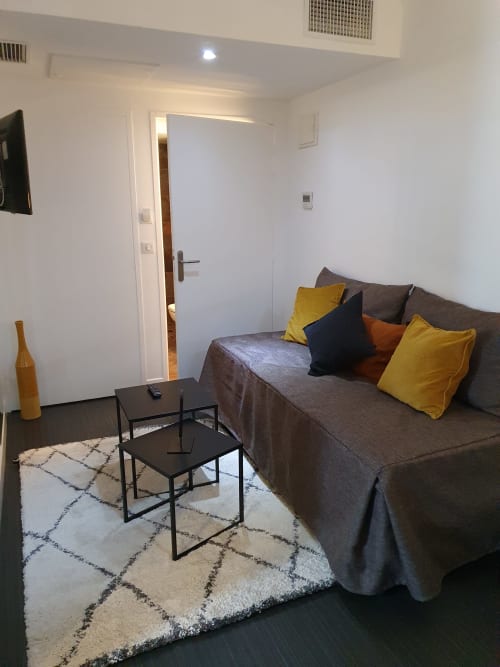 Suite junior moderne à l'Hôtel Au Bon Coin à Biarritz avec canapé gris, coussins jaunes, tables d'appoint noires et porte avec vue sur la ville