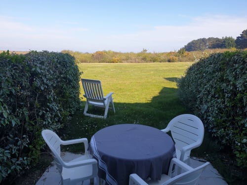 Coin détente du jardin avec table ronde, chaises blanches et haies de lierre, vue sur les champs verts de Locmaria, Belle Île