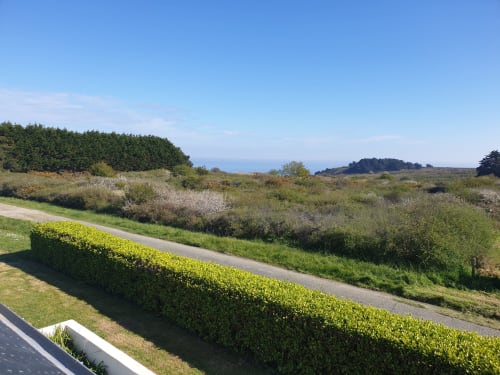 Vue sur le jardin depuis l'appartement de Locmaria, avec paysage côtier, haies, champs verdoyants et horizon marin sous ciel bleu