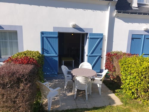 Patio ensoleillé de l'appartement Loc'Maria Belle Île avec mobilier blanc, haies colorées, volets bleus et façade cottage blanche