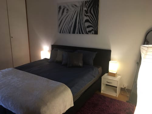 Modern bedroom at Au P'tit Bonheur B&B with black bed, dark bedding, white nightstand with lamp, zebra artwork, and burgundy