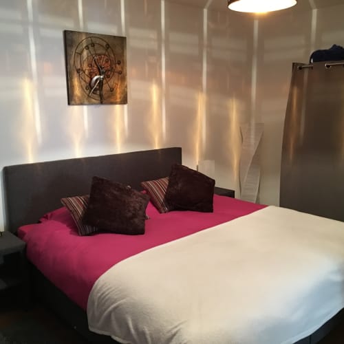 Modern bedroom at Au P'tit Bonheur cottage with pink bedding, black pillows, decorative wall clock, and garden views through
