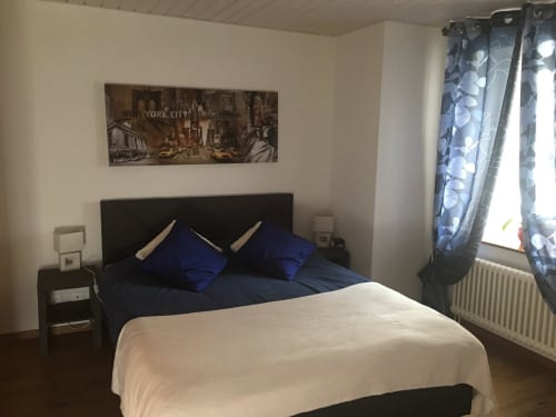 Cozy bedroom at Au P'tit Bonheur with blue pillows, white blanket, black headboard, and New York City artwork above bed