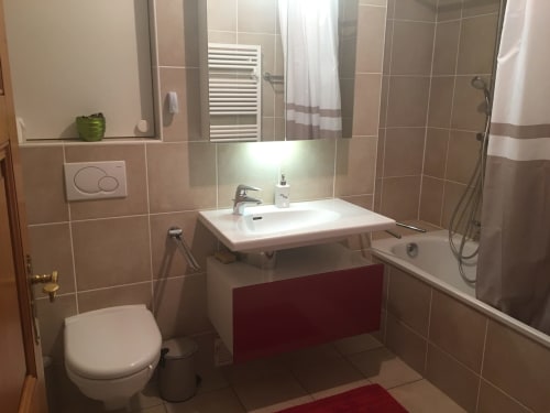 Modern bathroom with beige tiles, white sink, toilet, bathtub with shower, and red accent cabinet at Au P'tit Bonheur B&B