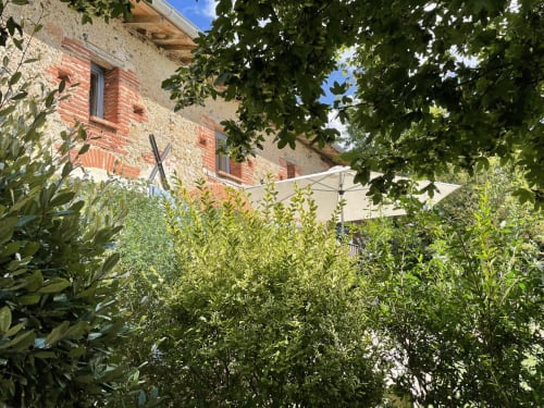 Cour verdoyante de la Maison Monsieur Léger B&B à Montaut, avec bâtisse en pierre et brique, parasol blanc et jardin luxuriant