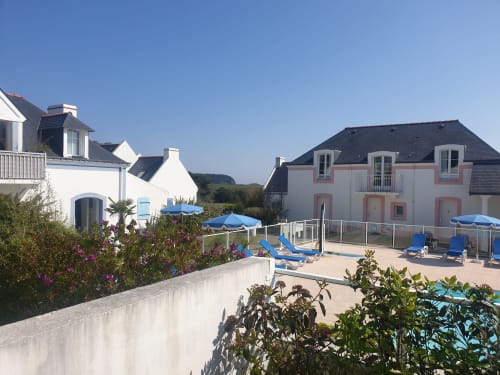 Espace piscine de Loc'Maria Belle Île-en-Mer avec parasols bleus, chaises longues et bâtiments résidentiels colorés en arrière-plan