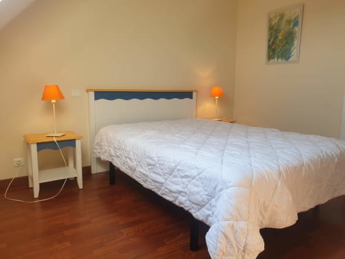 Chambre lumineuse dans un appartement de Belle Île en Mer avec lit blanc matelassé, tête de lit bleu marine, lampes orange et parquet