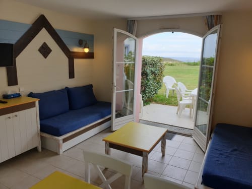 Appartement accueillant à Loc'Maria Belle Île avec sièges bleus, table à manger et portes-fenêtres donnant sur une terrasse avec vue mer et espaces verts
