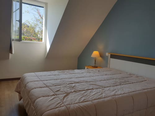 Chambre lumineuse sous les toits avec plafond mansardé, lit blanc douillet, lampe de chevet et fenêtre donnant sur le jardin à Belle Île en