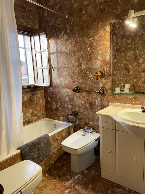 Baño con paredes de azulejos de mármol marrón, tocador con lavabo blanco, inodoro, bañera, espejo y ventana en Chalets Junto A Puy Du Fou