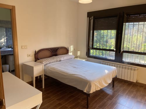Dormitorio luminoso en la cabaña de Argés con cama de madera, ropa de cama blanca, vistas a las montañas por grandes ventanales y comodidades modernas