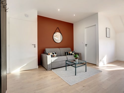 Salon moderne avec canapé gris, mur d'accent ocre et miroir rond à la Résidence L'Hermitière, Loire-Atlantique