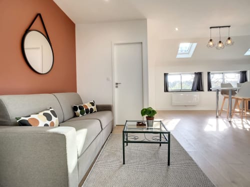 Salon moderne de l'appartement Résidence L'Hermitière avec canapé gris, mur d'accent orange, miroir rond et vue sur le jardin