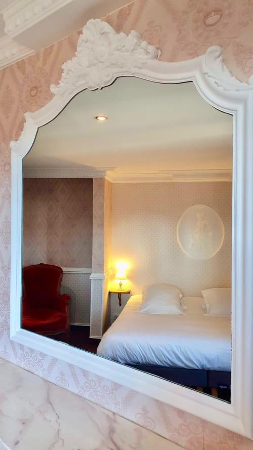 Chambre d'hôtel Les Cabines reflétée dans un miroir orné à cadre blanc, montrant le lit, la lampe de chevet et la chaise rouge