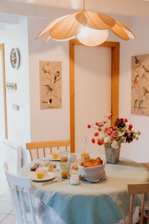 Petit-déjeuner lumineux aux Gites du Bulz avec lampe suspendue en bois, fleurs fraiches, croissants et œuvre d'art ornithologique