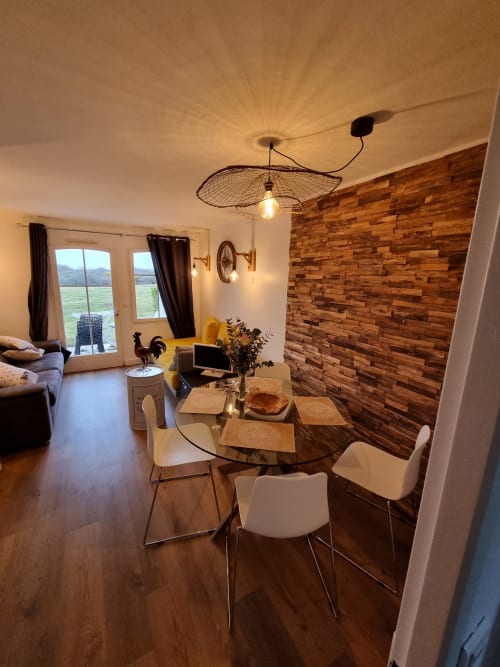 Salle à manger contemporaine dans un appartement de Belle-Île-en-Mer avec table ronde en verre, chaises blanches, mur en bois et vue sur la mer campagnarde