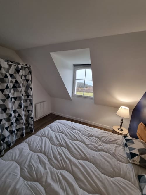Chambre mansardée lumineuse avec lucarne surplombant le paysage de Belle Île, rideaux géométriques et vue sur la mer côtière