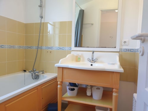 Salle de bain de l'appartement Locmaria : carrelage jaune, meuble vasque en bois, miroir et baignoire avec douche.