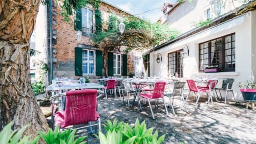 Cour charmante de La Maison De Fleurat à Domeyrot avec chaises rouges, tables de salle à manger et bâtiments en pierre couverts de lierre.
