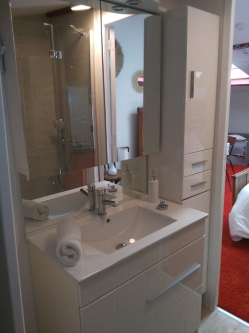 Vue sur l'espace vasque de la salle de bain attenante à la chambre familiale Soleil à La Chabanise. Un meuble sous-vasque blanc laqué avec tiroirs, une colonne de rangement, un grand miroir éclairé et une serviette enroulée. Le reflet montre la douche et l'ouverture donne sur la chambre.