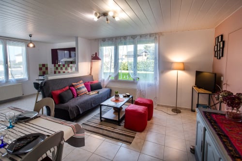 Séjour ouvert à Au Gite d'Isigny avec canapé gris, poufs rouges, kitchenette et vue sur balcon