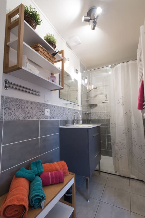 Modern bathroom at Au Gite d'Isigny with dark tiles, wooden shelving, colorful towels, and glass shower enclosure