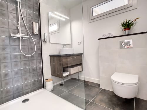 Salle de bain moderne à la Résidence L'Hermitière avec douche à l'italienne, carrelage gris foncé, vasque blanche et toilettes