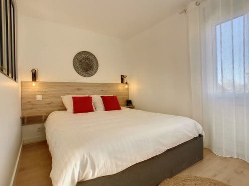 Chambre moderne à la Résidence L'Hermitière avec tête de lit en bois, literie blanche, oreillers rouges et vue sur le jardin