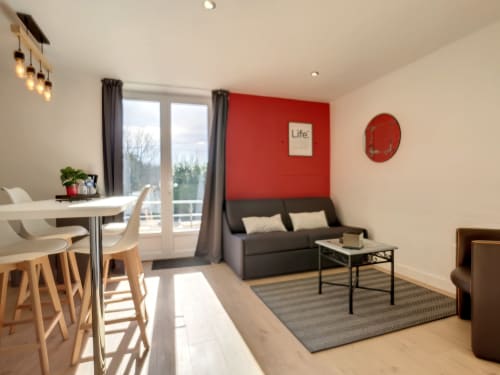 Studio moderne à la Résidence L'Hermitière avec mur d'accent rouge, coin repas, canapé et vue sur le jardin depuis le balcon